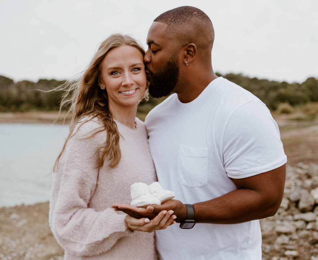 Husband kisses wife while she holds baby booties at their fun pregnancy announcement photoshoot. 