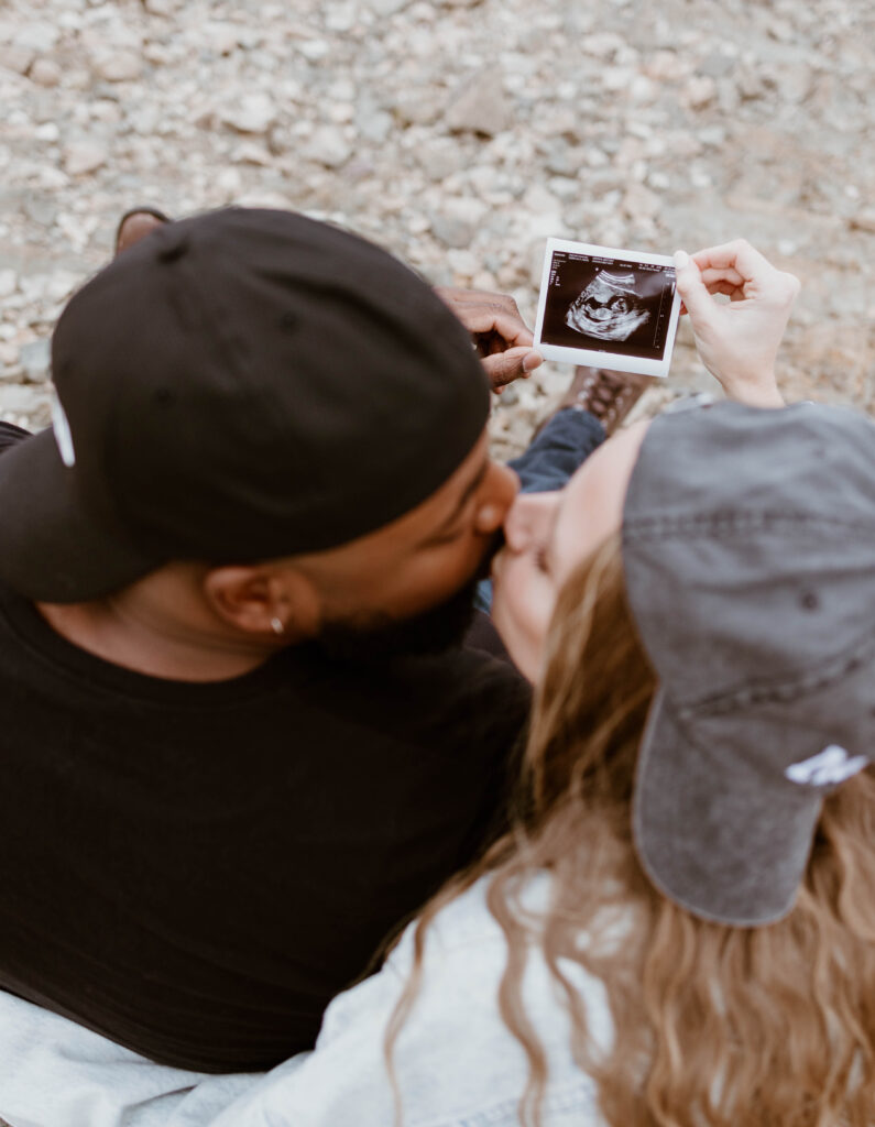 Husband and wife kiss while holding ultrasound at fun pregnancy announcement photoshoot