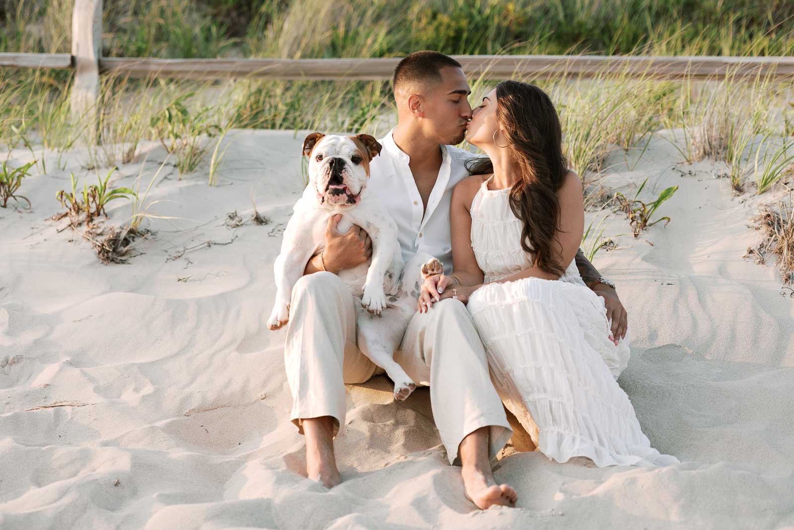 Engaged couple kisses on the beach with bulldog